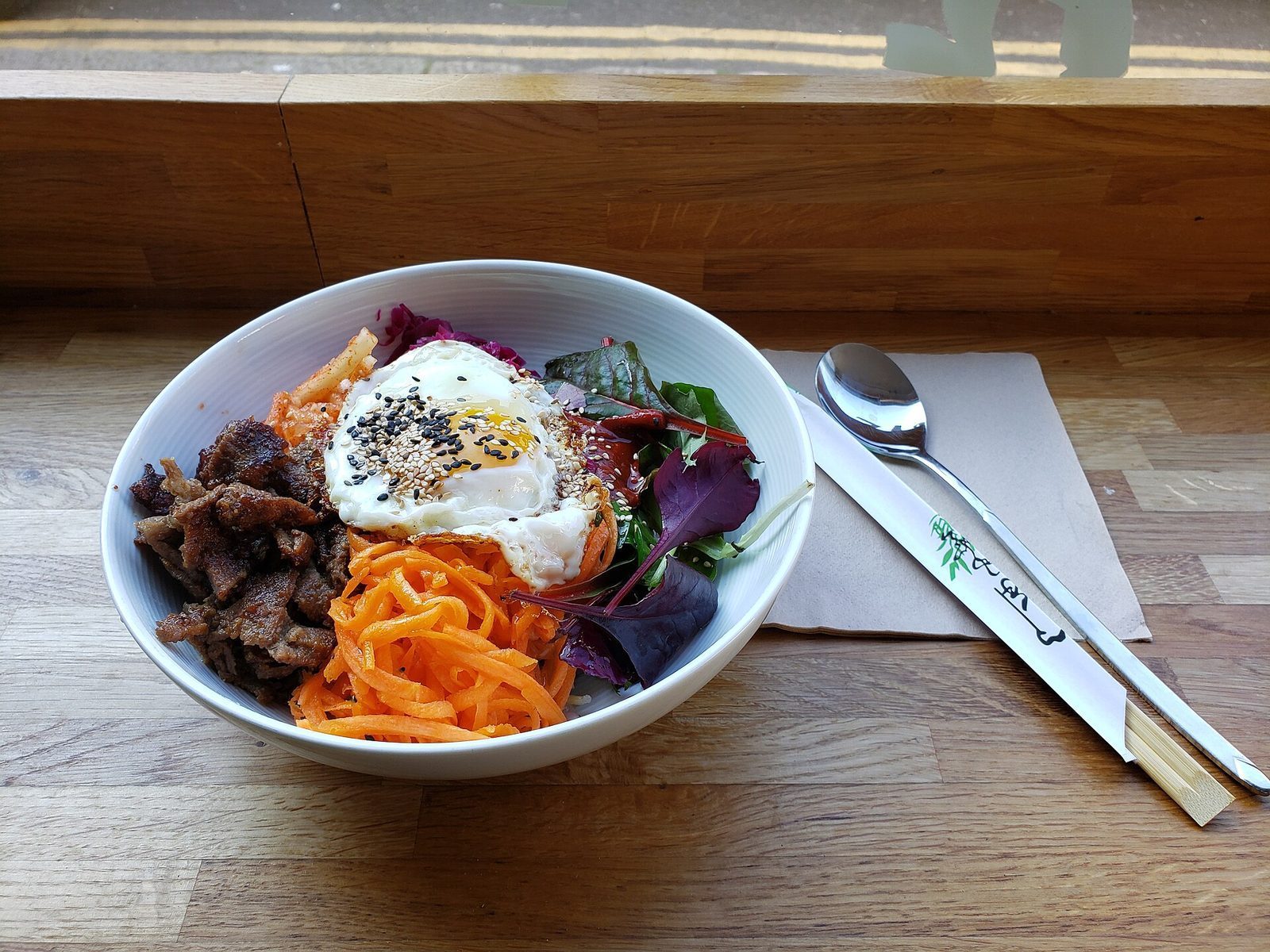 Bibimbap i hvit skål med speilegg drysset med sesam, marinert biff, revet gulrot og friske salatblader ved et vindu