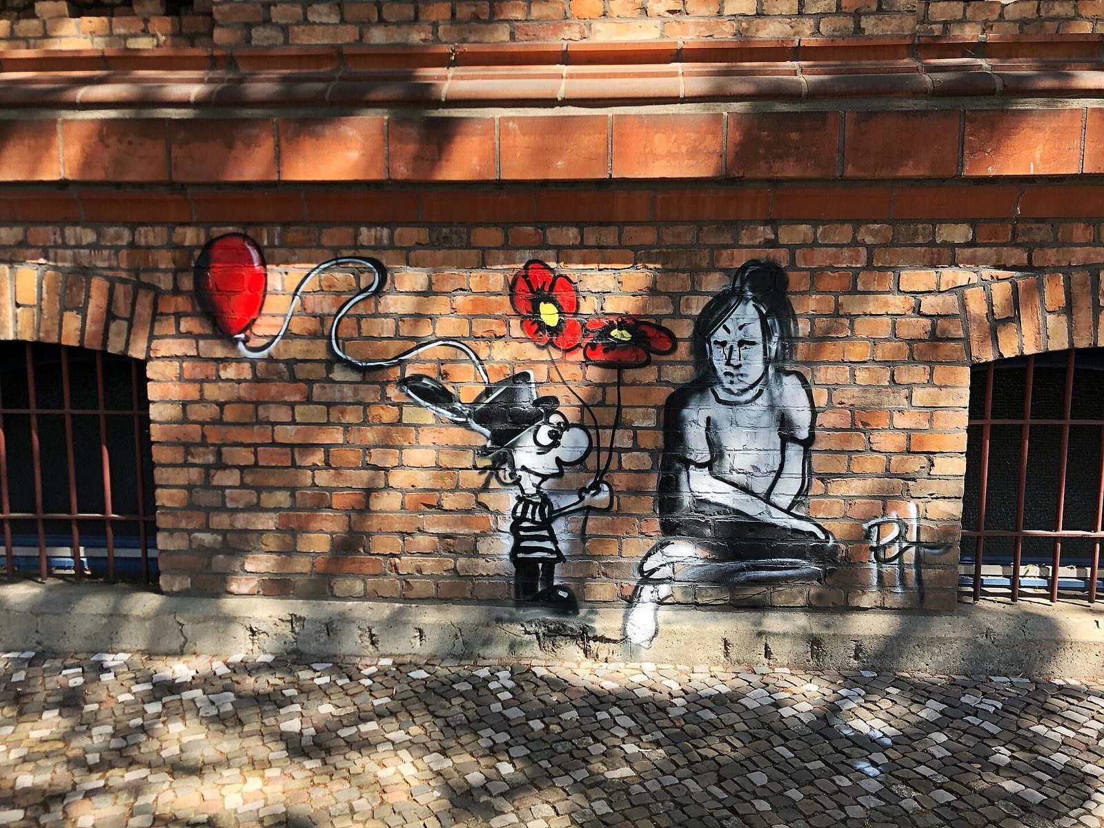 Sjablongmaleri pa teglvegg i Wrangelstrasse: en guttefigur med rod ballong og blomst ved siden av en sittende jente