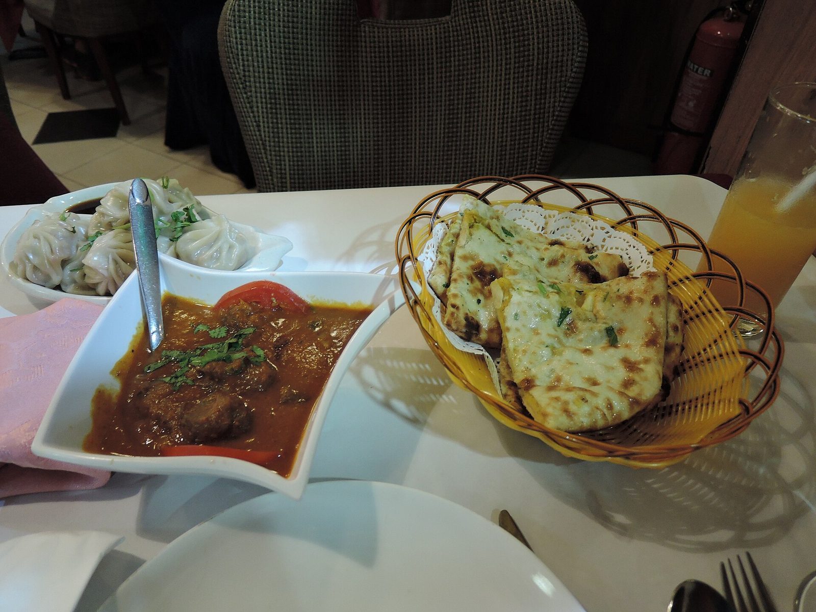 Kjøttkarri i skål ved siden av en kurv med nystekt naanbrød og en porsjon momos på et restaurantbord