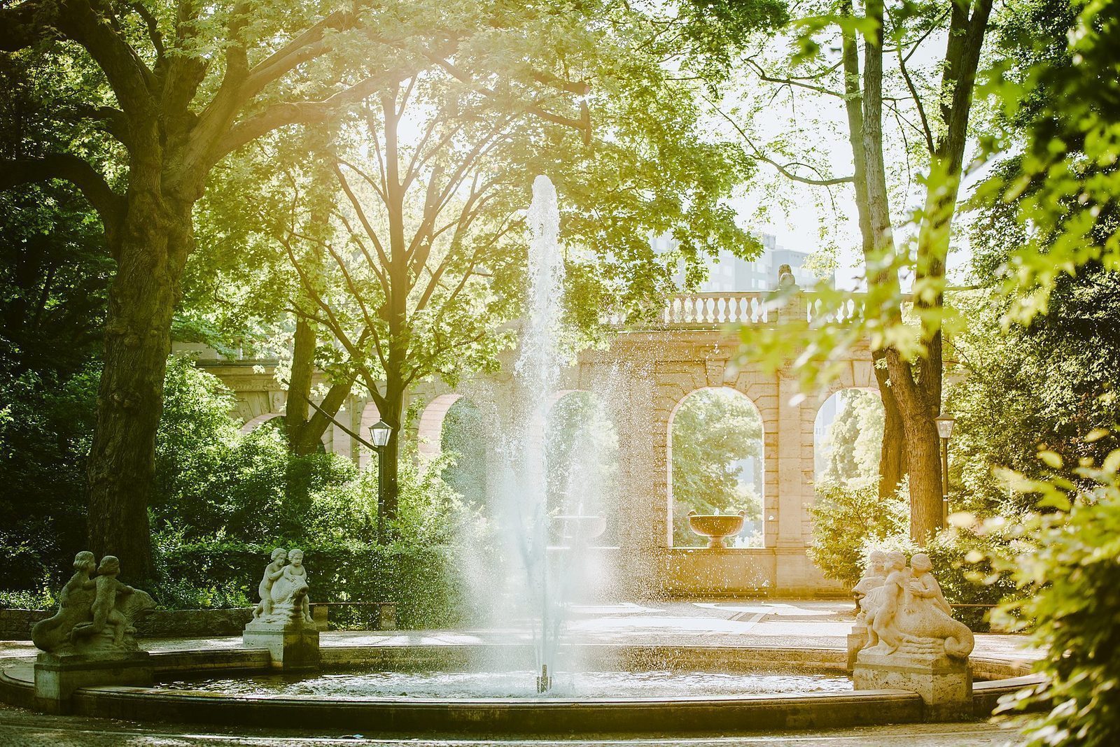 En høy vannstråle i motlys ved Märchenbrunnen, rammet inn av søylegangen og fontenefigurene