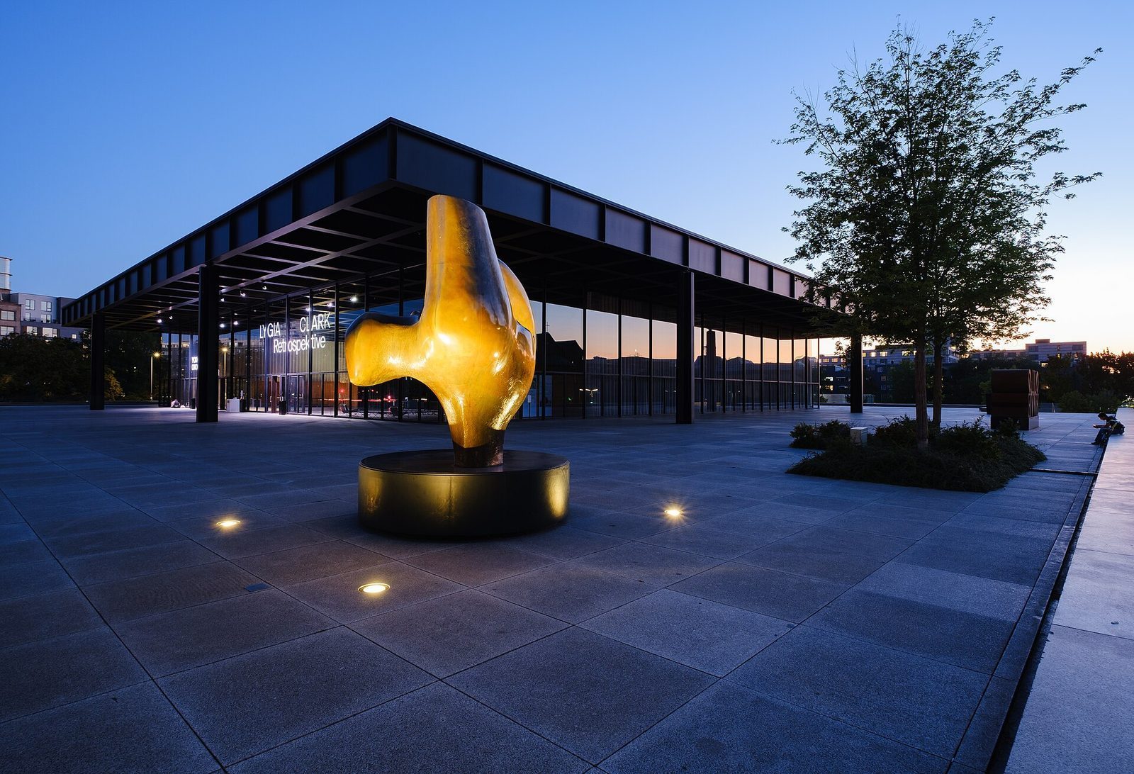 En stor gyllen, biomorf skulptur på granittplassen foran Neue Nationalgalerie i blåtimen, med glasshallen opplyst bak