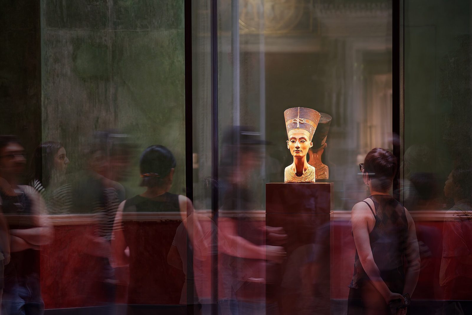 Nefertiti-bysten opplyst i glassmonteren i Neues Museum, med besøkende speilet i glasset