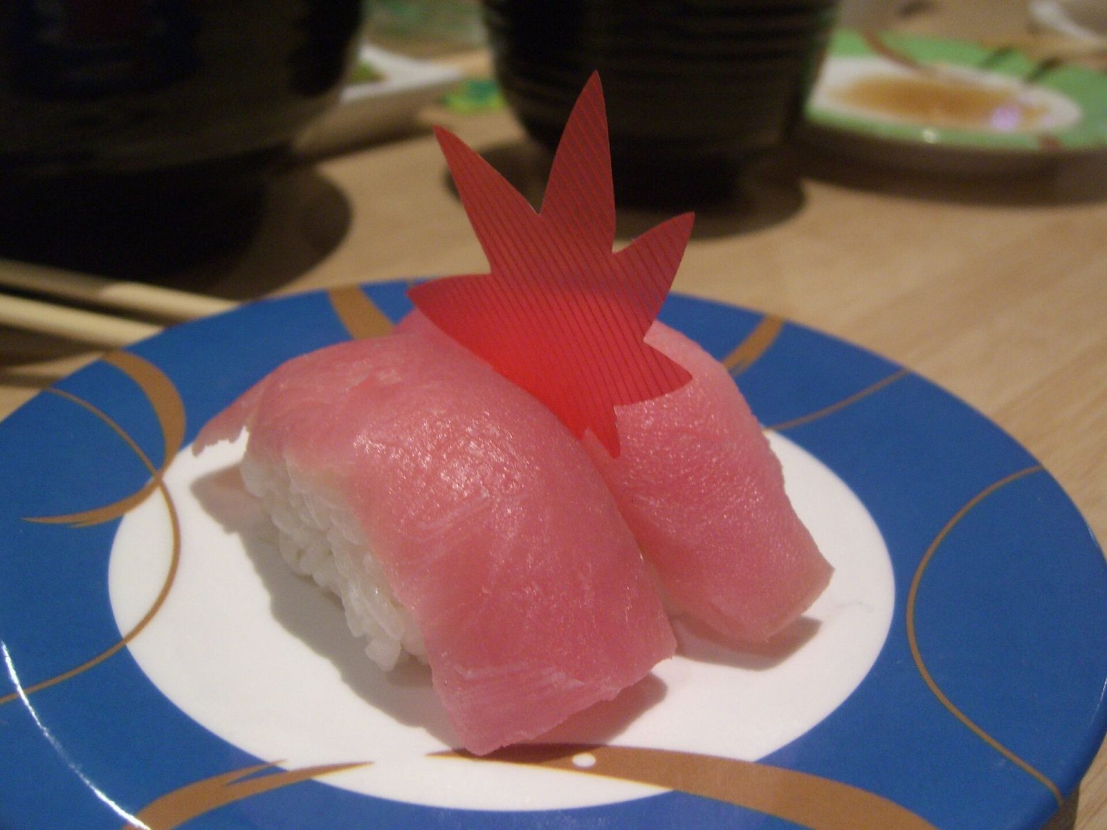 To biter tunfisk-nigiri på en blå tallerken med gullkant og en rød dekorasjon i bakgrunnen