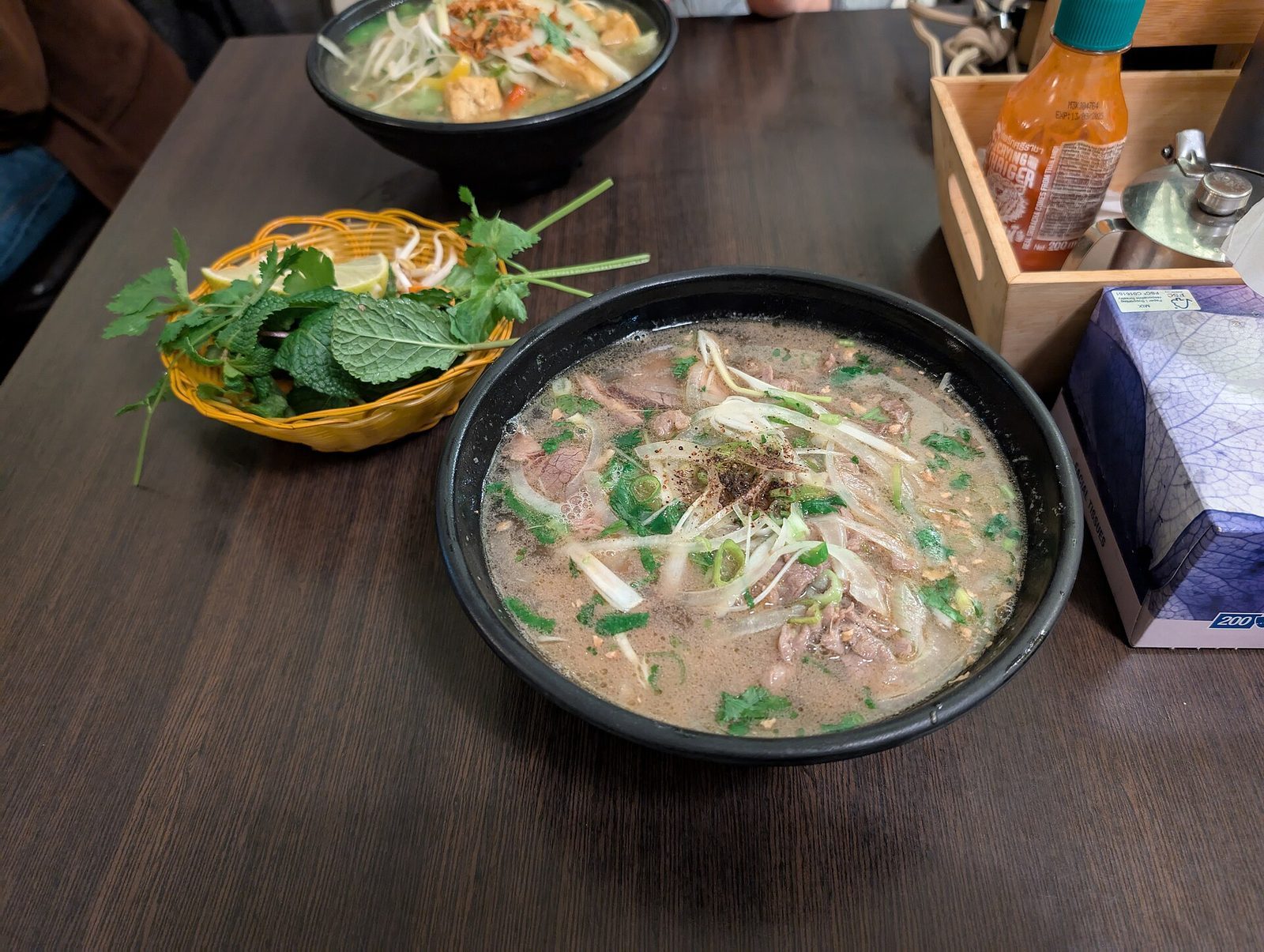 Skål med phở bò på restaurantbord, med kurv med friske urter og sriracha ved siden