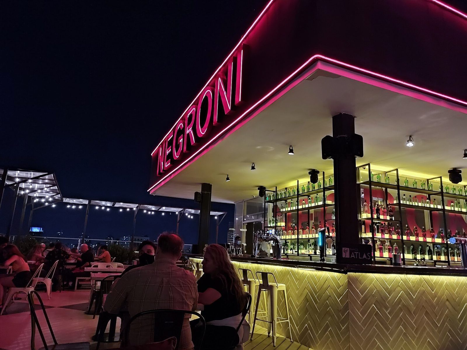 Takbar om kvelden med rosa NEGRONI-neonskilt, opplyst bardisk og gjester ved bordene