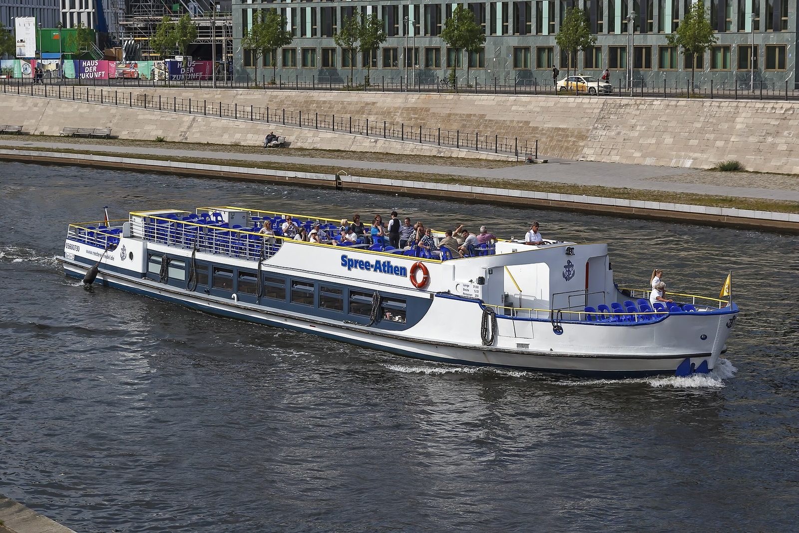 Utfluktsbåten «Spree-Athen» på elva Spree med passasjerer på åpent soldekk, langs en moderne bebygd elvebredd