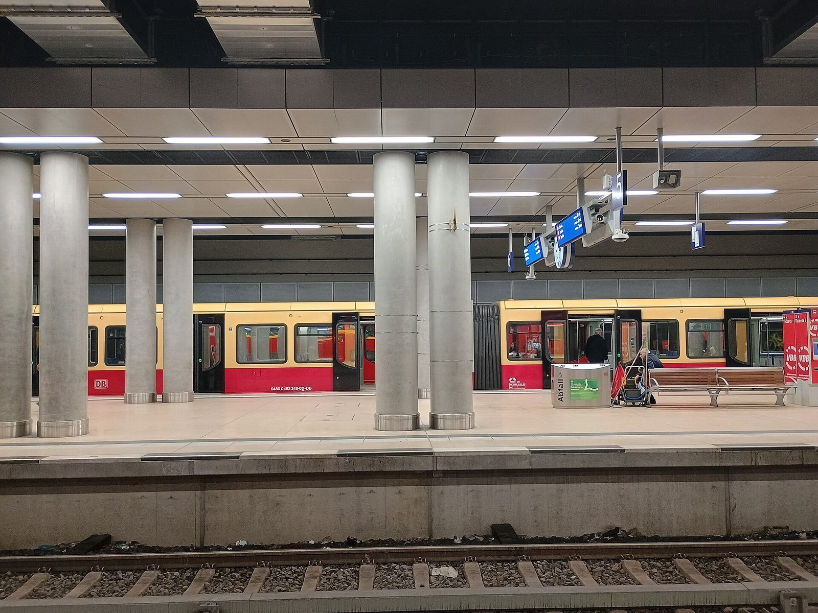 Perrongen pa jernbanestasjonen under BER, med et rod-gult S-Bahn-tog og en rod VBB-billettautomat