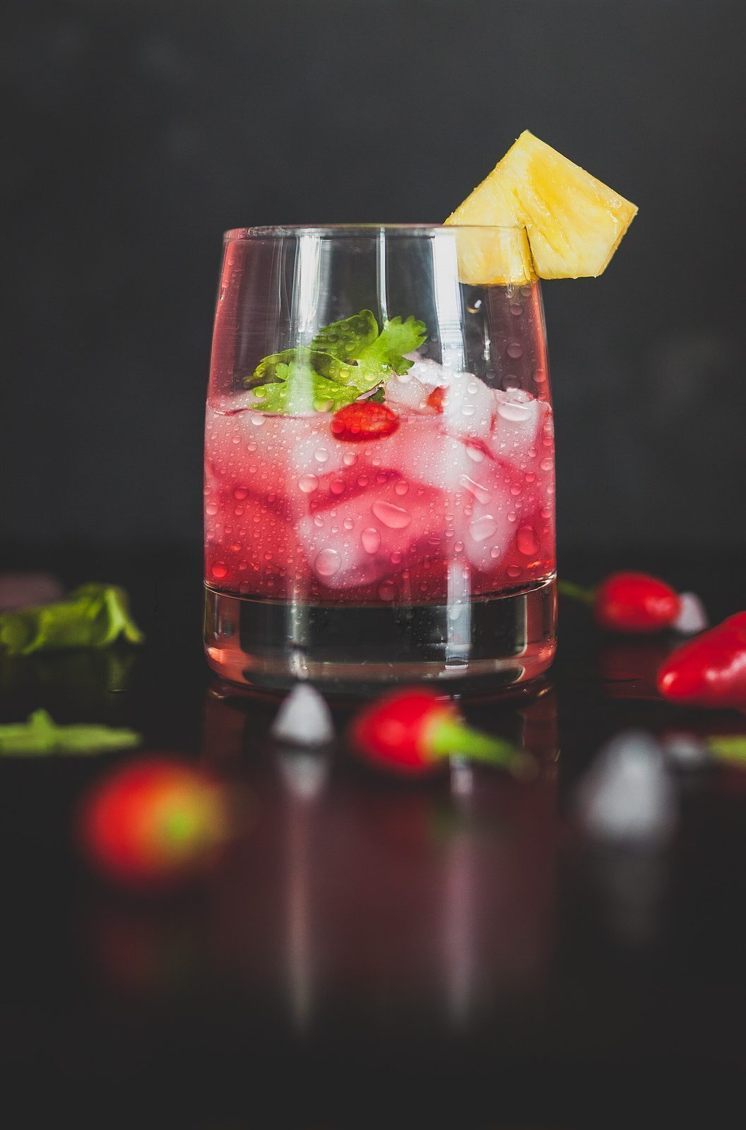 Rosa cocktail i lavt glass med ananasbit og korianderblad, mot mørk bakgrunn
