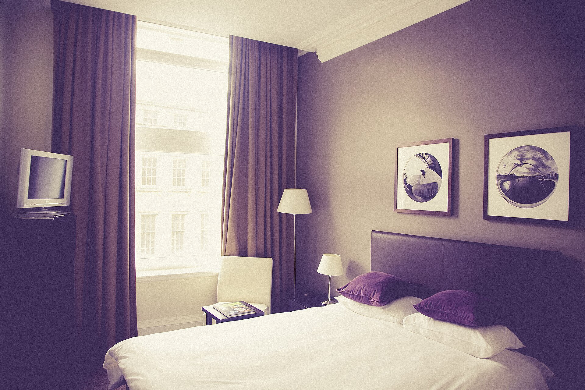 Hotellrom med lilla vegger, dobbeltseng og innrammede bilder over sengegavlen